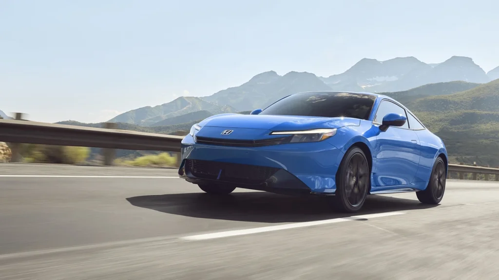 A blue Honda Prelude 2026 driving on a highway with mountains in the background, showing off its sporty and aerodynamic front design.