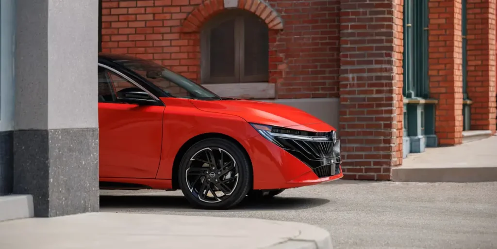 a red 2026 Nissan Sentra is shown in a close up profile shot, highlighting its redesigned front end, new headlights, and intricate wheel design