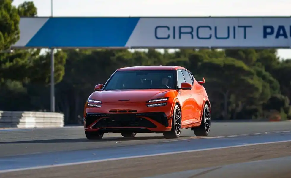 Rear angle of the 2027 Genesis GV60 Magma EV on a race track, highlighting its sporty rear spoiler and widened stance.